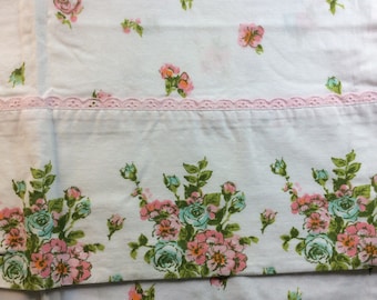 May include: White floral bed sheet with a pink lace trim. The sheet features a pattern of pink and teal roses with green leaves. The roses are scattered across the sheet and clustered at the bottom.