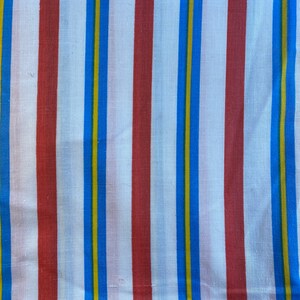 May include: Close-up of a fabric with vertical stripes. The stripes alternate between white, red, blue, and yellow. The fabric has a slightly wrinkled texture, suggesting it is made of a natural material like cotton or linen.