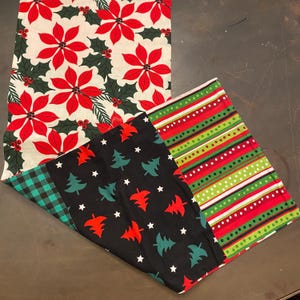 May include: A collection of Christmas-themed fabric table runners. One features red poinsettias and holly on a cream background. Another has a black background with red trees, green trees, and white stars. The third has red and green stripes.