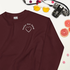 Pode incluir: Camisola de cor bordô com um coração bordado branco e o texto "Tachy Girl Gang" no peito esquerdo.