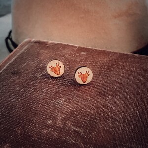 Puede incluir: Un par de pendientes de madera con un diseño de cabeza de ciervo. Los pendientes son de color marrón claro y tienen un acabado de madera natural.