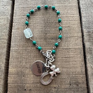 May include: A green beaded bracelet with a silver charm that says "Courage" and features a horseshoe, a shamrock, and a clear crystal.