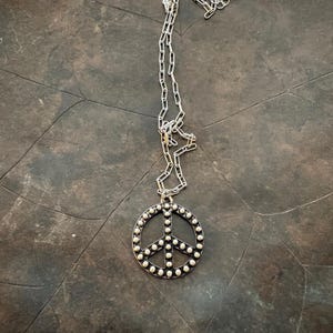 May include: Silver-toned peace sign pendant necklace. The pendant is a circle with the peace symbol in the center, surrounded by small, round embellishments. The chain is a delicate link style. The necklace is displayed on a dark, textured surface.