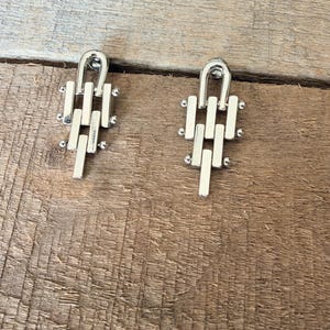 May include: A pair of silver-toned earrings featuring a vertical, multi-layered chain link design. Each earring consists of a top U-shaped loop connected to three descending rectangular bars, accented with small spherical beads at the joints.