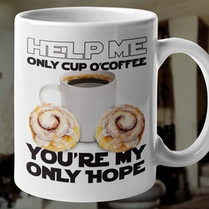 May include: White ceramic coffee mug with a black and white graphic that says "Help me, only cup o' coffee, you're my only hope". The mug has a coffee cup with coffee and two cinnamon rolls on it.