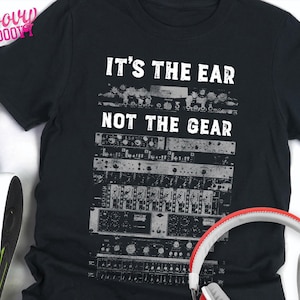May include: A black short-sleeved t-shirt featuring a white graphic print of stacked audio recording equipment and the text "IT'S THE EAR NOT THE GEAR" in a distressed font. The design is centered on the chest of the garment.