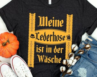 Puede incluir: Una camiseta gris carbón oscuro con un estampado gráfico amarillo que incluye el texto "Meine Lederhose ist in der Wäsche", flanqueado por bordes decorativos verticales, un icono de sombrero y un icono de acordeón.