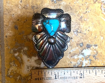 May include: An ornate silver brooch featuring a turquoise stone. The brooch has a detailed, symmetrical design with a heart-shaped turquoise stone at its centre. The brooch is approximately 5 cm in length.