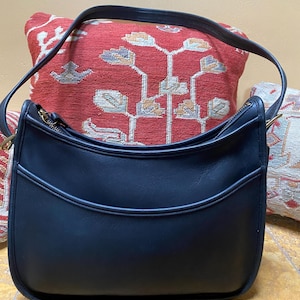May include: A black leather shoulder bag with a curved design. The bag has a top zipper closure and a long strap. The bag is set against a red patterned pillow.