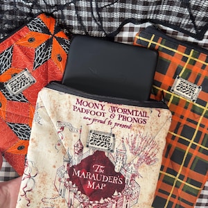 May include: Three fabric sleeves for electronic devices. One features a beige map print with text reading Moony, Wormtail, Padfoot & Prongs and The Marauder's Map. Another has an orange diamond pattern with spiders. The third has an orange plaid.