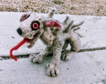 May include: A gray and white felt creature with a long red tongue sticking out. The creature has a large, red eye and a menacing expression.