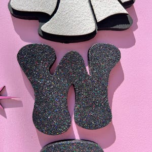 May include: Close-up of decorative glittery letters and shapes on a pink background. The letter "W" is in dark gray glitter. A white glittery shape with black trim is at the top. The overall theme is a craft or party decoration.