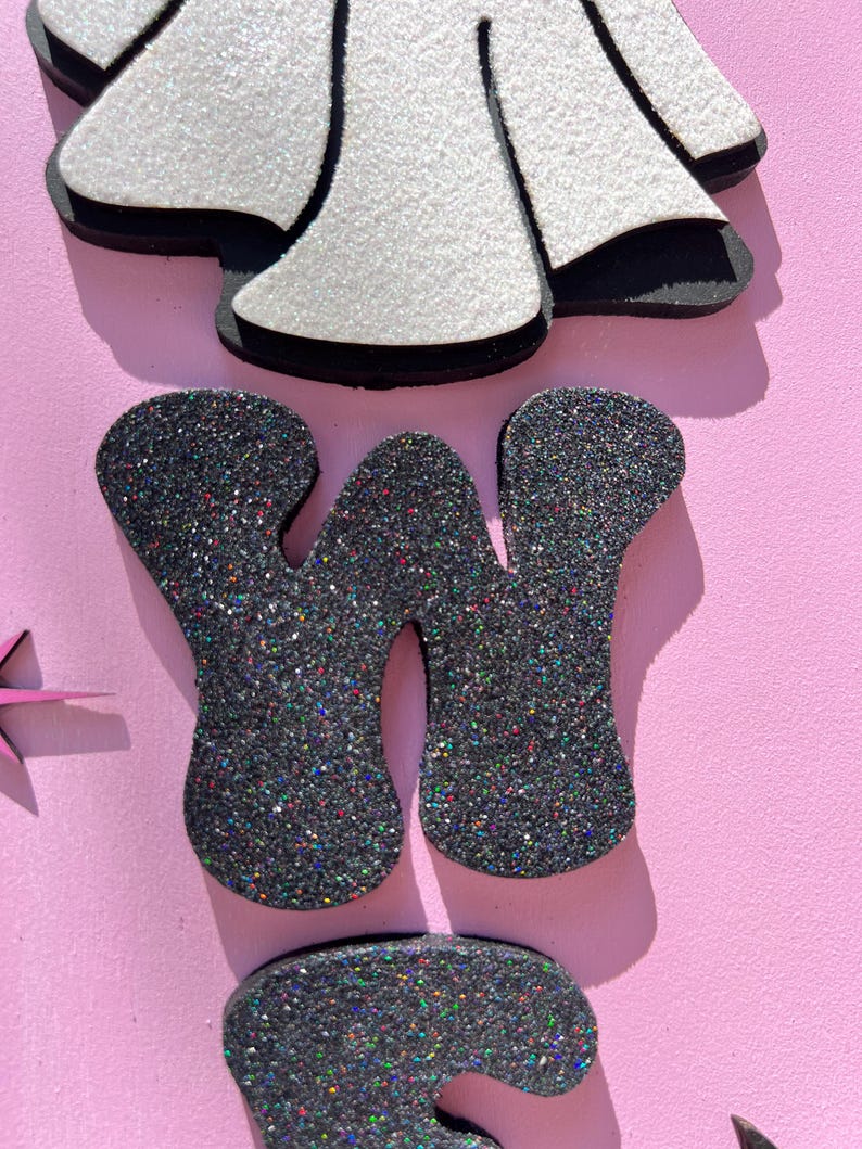May include: Close-up of decorative glittery letters and shapes on a pink background. The letter "W" is in dark gray glitter. A white glittery shape with black trim is at the top. The overall theme is a craft or party decoration.