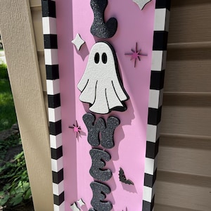 May include: A pink Halloween sign with the word "Halloween" in black glitter letters. A white ghost and star accents are also featured. The sign has a black and white striped border.