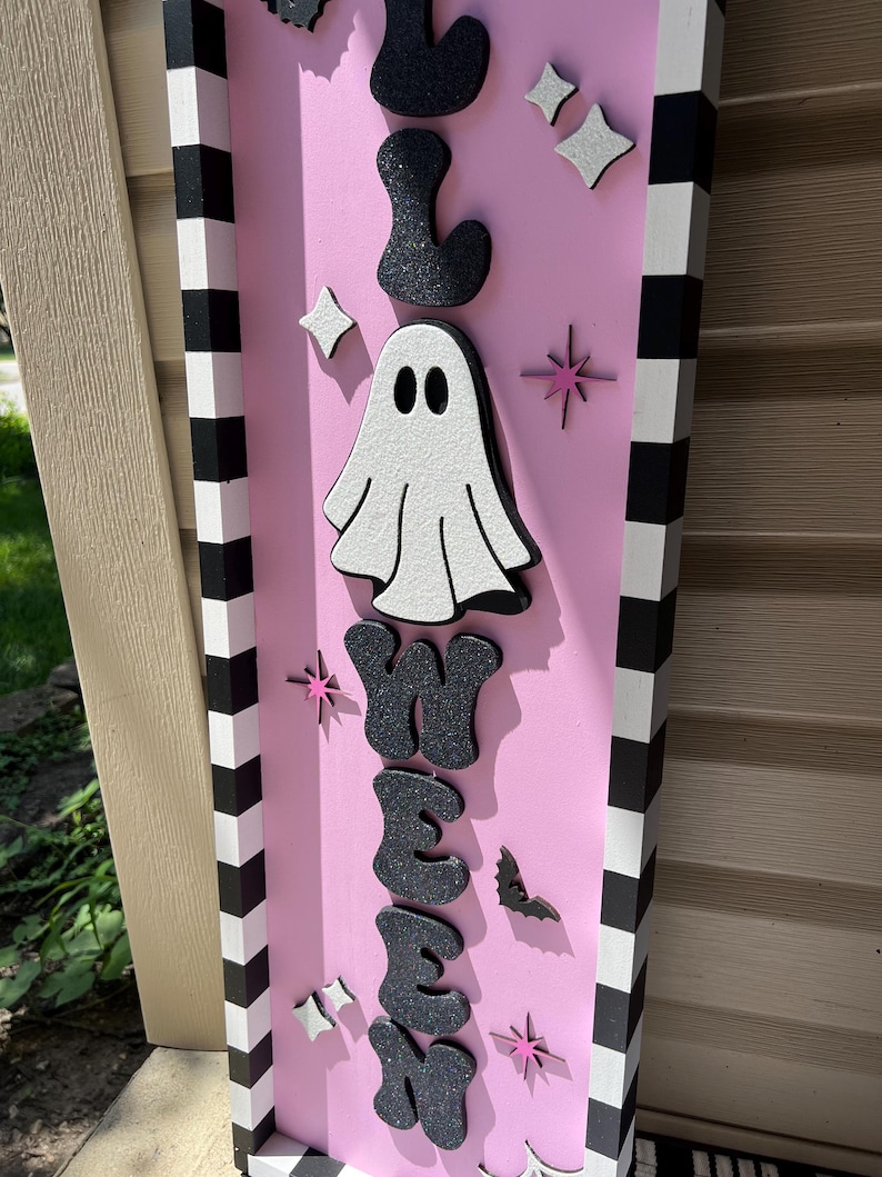 May include: A pink Halloween sign with the word "Halloween" in black glitter letters. A white ghost and star accents are also featured. The sign has a black and white striped border.