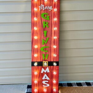May include: A tall rectangular wooden sign with a red wood grain finish, featuring the text Merry GRINCH MAS in white and green lettering. The sign is illuminated by two vertical rows of small round light bulbs and includes a black belt detail.