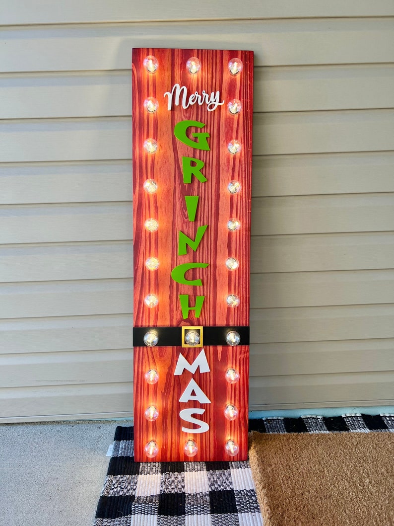 May include: A tall rectangular wooden sign with a red wood grain finish, featuring the text Merry GRINCH MAS in white and green lettering. The sign is illuminated by two vertical rows of small round light bulbs and includes a black belt detail.