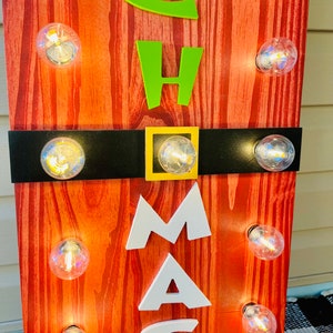 May include: A vertical wooden sign with a red wood grain finish featuring the letters C, H, A, M, A, S in lime green and white. A horizontal black band with a gold square buckle crosses the center. Ten clear round light bulbs are attached.