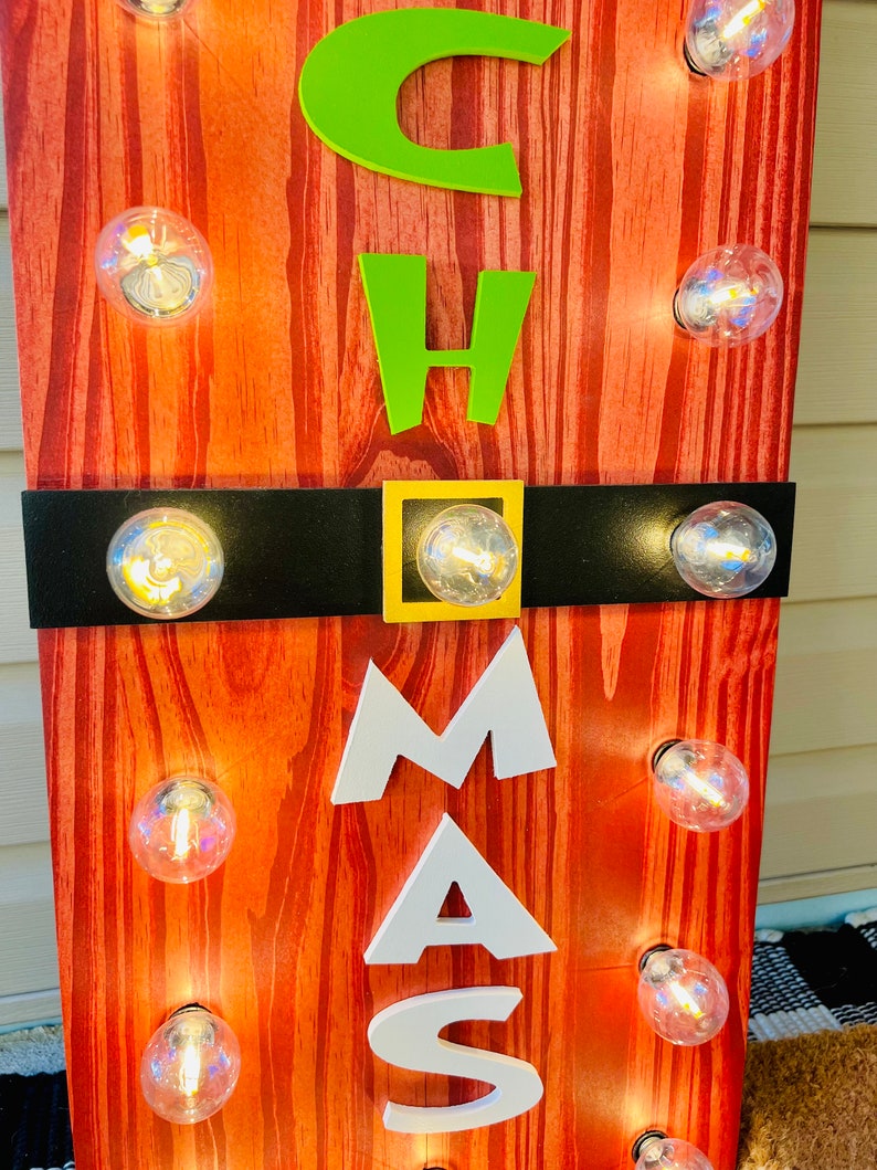 May include: A vertical wooden sign with a red wood grain finish featuring the letters C, H, A, M, A, S in lime green and white. A horizontal black band with a gold square buckle crosses the center. Ten clear round light bulbs are attached.