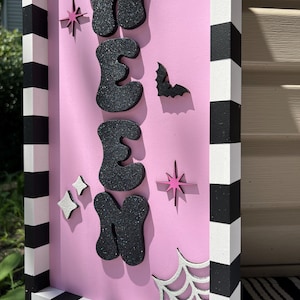 May include: A pink Halloween sign with the word "WEEN" in black glitter letters. The sign features a bat, stars, and a spiderweb. The frame is black and white striped.