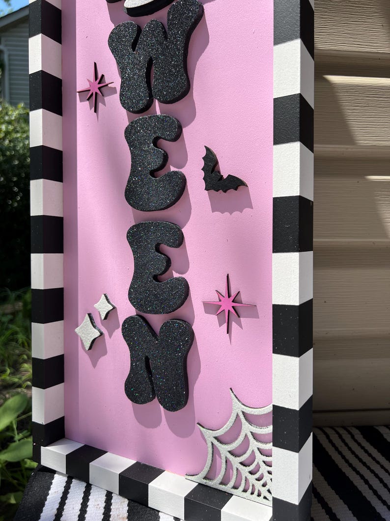 May include: A pink Halloween sign with the word "WEEN" in black glitter letters. The sign features a bat, stars, and a spiderweb. The frame is black and white striped.