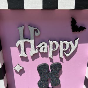 May include: A pink Halloween-themed box with the word "Happy" in white and silver letters. The box is decorated with a black bat, starbursts, and glittery black shapes. The top and sides have a black and white checkered pattern.