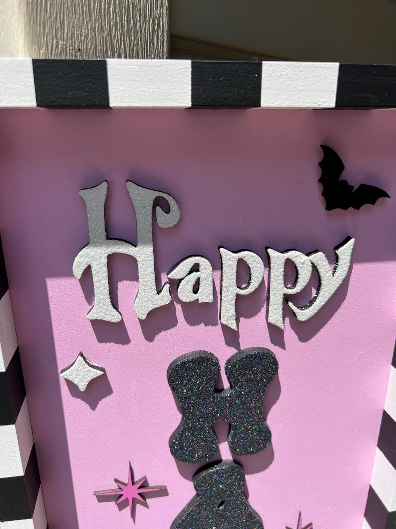 May include: A pink Halloween-themed box with the word "Happy" in white and silver letters. The box is decorated with a black bat, starbursts, and glittery black shapes. The top and sides have a black and white checkered pattern.