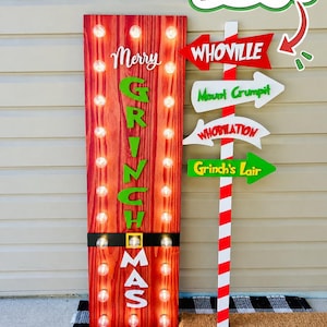 May include: A tall rectangular red wood-grain sign with vertical rows of light bulbs, featuring the text Merry Grinchmas in green and white, paired with a red and white striped pole sign pointing to Whoville, Mount Crumpit, Whobilation, and Grinch's Lair.