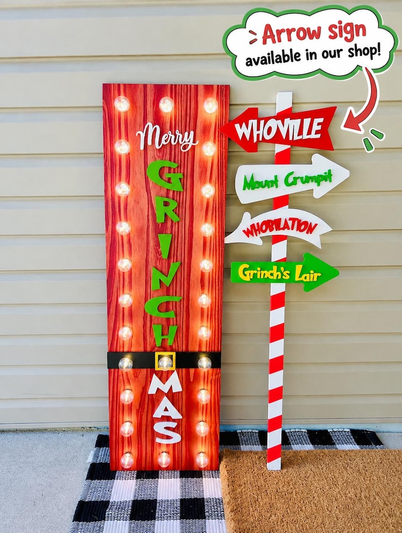 May include: A tall rectangular red wood-grain sign with vertical rows of light bulbs, featuring the text Merry Grinchmas in green and white, paired with a red and white striped pole sign pointing to Whoville, Mount Crumpit, Whobilation, and Grinch's Lair.