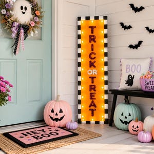 Puede incluir: Un letrero luminoso rectangular alto con un borde a cuadros blancos y negros y un centro naranja que muestra el texto TRICK OR TREAT en letras negras. El letrero est&aacute; rodeado de decoraciones de Halloween, calabazas y una corona.