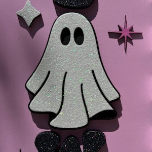 May include: A glittery white ghost with black eyes and a black outline, surrounded by star and crown shapes, against a pink background. The Halloween-themed design features a sparkly, textured appearance.