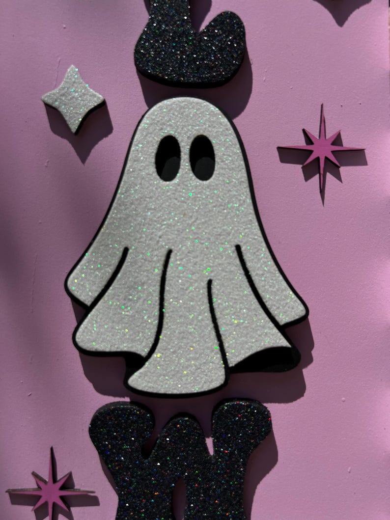 May include: A glittery white ghost with black eyes and a black outline, surrounded by star and crown shapes, against a pink background. The Halloween-themed design features a sparkly, textured appearance.