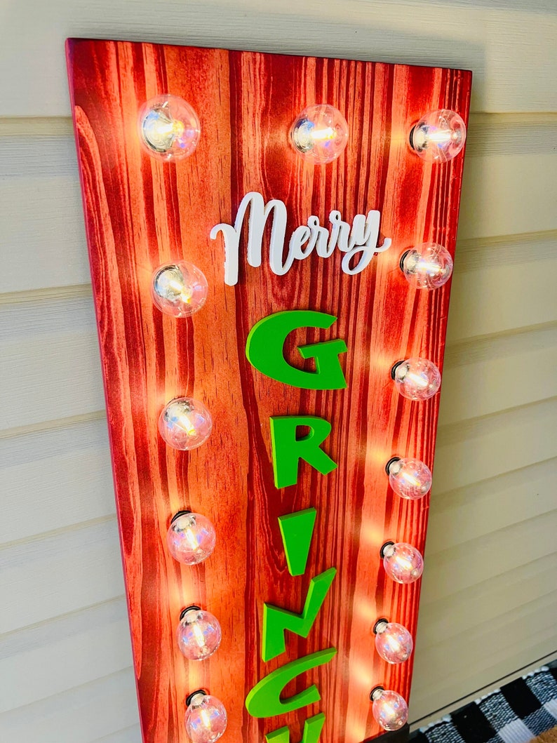May include: A vertical wooden sign with a red wood grain finish featuring the white script text Merry and the green block letter text GRINCH. The sign is illuminated by ten round clear light bulbs arranged in two columns along the sides.