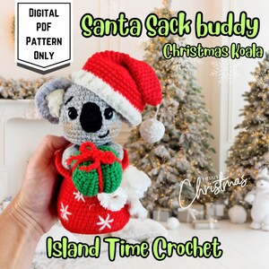 May include: A crocheted Christmas Koala wearing a Santa hat and holding a green gift in a red sack. The sack is decorated with white snowflakes. Text on the image reads "Santa Sack Buddy", "Christmas Koala", "Digital PDF Pattern Only", "Island Time Crochet", and "Merry Christmas".