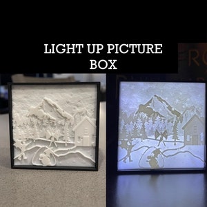 May include: A white light-up picture box with a winter scene. The scene features a snowy mountain range, trees, a house, and two figures. The box is lit up, making the scene glow.