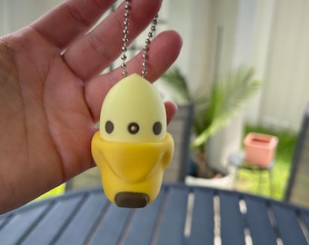 May include: A yellow and white keychain shaped like a cartoon chick with a silver chain. The chick has two black eyes and a brown beak.