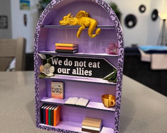 Peut inclure: Une petite bibliothèque miniature violette avec une figurine de dragon dorée, une pancarte qui dit "We do not eat our allies", et plusieurs livres. La bibliothèque a une bordure décorative noire et blanche.