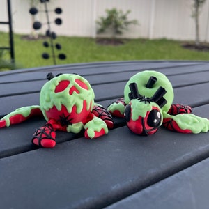 Puede incluir: Dos figuras de ara&ntilde;as con tem&aacute;tica de Halloween. Las ara&ntilde;as tienen cuerpos rojos con caras y patas verdes goteantes en forma de calavera. Tambi&eacute;n se ven telara&ntilde;as negras y peque&ntilde;as ara&ntilde;as negras. Las figuras est&aacute;n sobre una superficie gris oscuro.