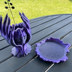 May include: A purple 3D printed decorative dish with a wavy edge and a smooth, round interior.  A matching purple 3D printed sculpture with a face and hands raised in a gesture of offering sits next to the dish.