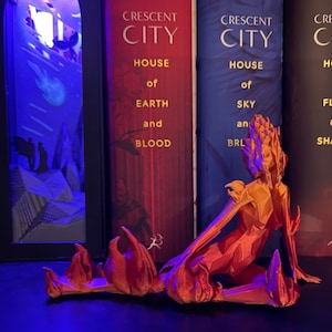 May include: A 3D printed figurine of a fire creature with a red and orange color scheme. The figurine is sitting on a shelf in front of three books with the titles "Crescent City House of Earth and Blood", "Crescent City House of Sky and Breath", and "Crescent City House of Flame and Shadow".