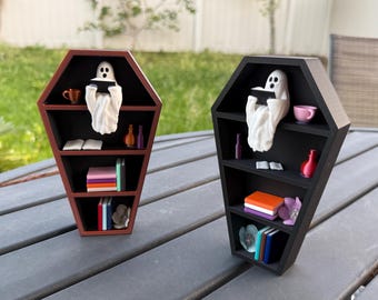 Puede incluir: Dos estanterías de libros en forma de ataúd negro con una figurita de fantasma leyendo un libro en cada estante. Las estanterías están llenas de libros, una taza de té y otros artículos decorativos. Una estantería está pintada de marrón y la otra de negro.