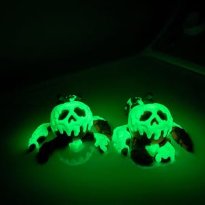 Puede incluir: Dos figuras brillantes en forma de calavera verde con ap&eacute;ndices negros. Las figuras tienen un aspecto goteante y derretido, y las calaveras tienen cuencas de ojos y una nariz huecas. El fondo oscuro enfatiza el brillo verde brillante.
