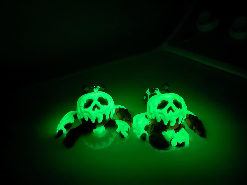 Puede incluir: Dos figuras brillantes en forma de calavera verde con ap&eacute;ndices negros. Las figuras tienen un aspecto goteante y derretido, y las calaveras tienen cuencas de ojos y una nariz huecas. El fondo oscuro enfatiza el brillo verde brillante.