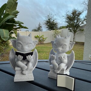 May include: Two gray gargoyle statues with wings and horns, each holding a small black book. The statues are seated on a gray surface, with a third book lying nearby. The background features a cloudy sky and green foliage.