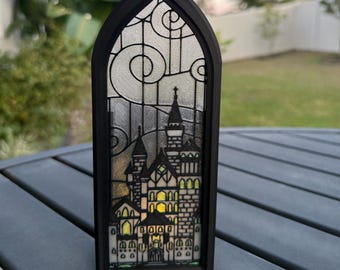 May include: A black framed, gothic-style window decoration featuring a castle design. The window has a stained-glass effect with a castle silhouette and decorative swirls. The castle has multiple towers and illuminated windows, creating a striking visual.