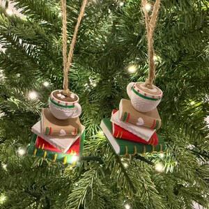 K&ouml;nnte beinhalten: Zwei Weihnachtsbaumschmuckst&uuml;cke. Jedes Ornament zeigt eine wei&szlig;e Tasse hei&szlig;e Schokolade mit gr&uuml;n-rotem Rand, auf einem Stapel B&uuml;cher. Die B&uuml;cher sind rot, wei&szlig; und gr&uuml;n mit goldenen Akzenten. Die Ornamente h&auml;ngen an Schnur.
