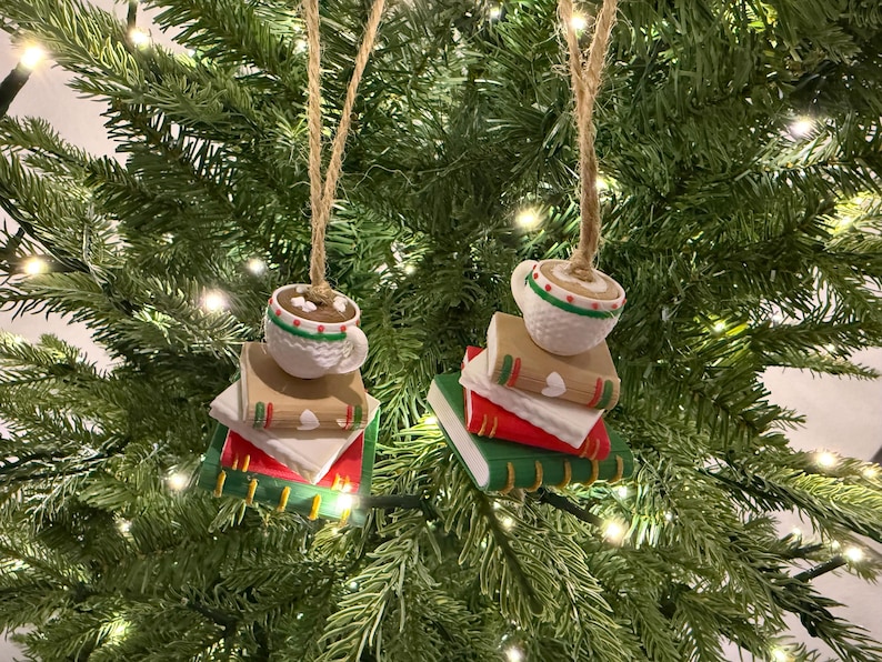 K&ouml;nnte beinhalten: Zwei Weihnachtsbaumschmuckst&uuml;cke. Jedes Ornament zeigt eine wei&szlig;e Tasse hei&szlig;e Schokolade mit gr&uuml;n-rotem Rand, auf einem Stapel B&uuml;cher. Die B&uuml;cher sind rot, wei&szlig; und gr&uuml;n mit goldenen Akzenten. Die Ornamente h&auml;ngen an Schnur.