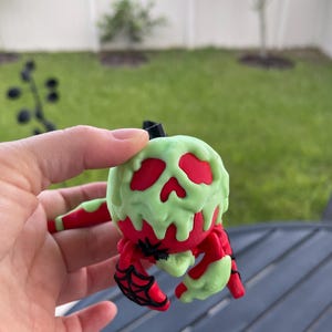 Puede incluir: Una figura de ara&ntilde;a roja y verde con tem&aacute;tica de Halloween. La ara&ntilde;a tiene un cuerpo rojo con detalles verdes goteando y detalles de telara&ntilde;a negros. La figura se sostiene en una mano sobre un fondo borroso al aire libre.
