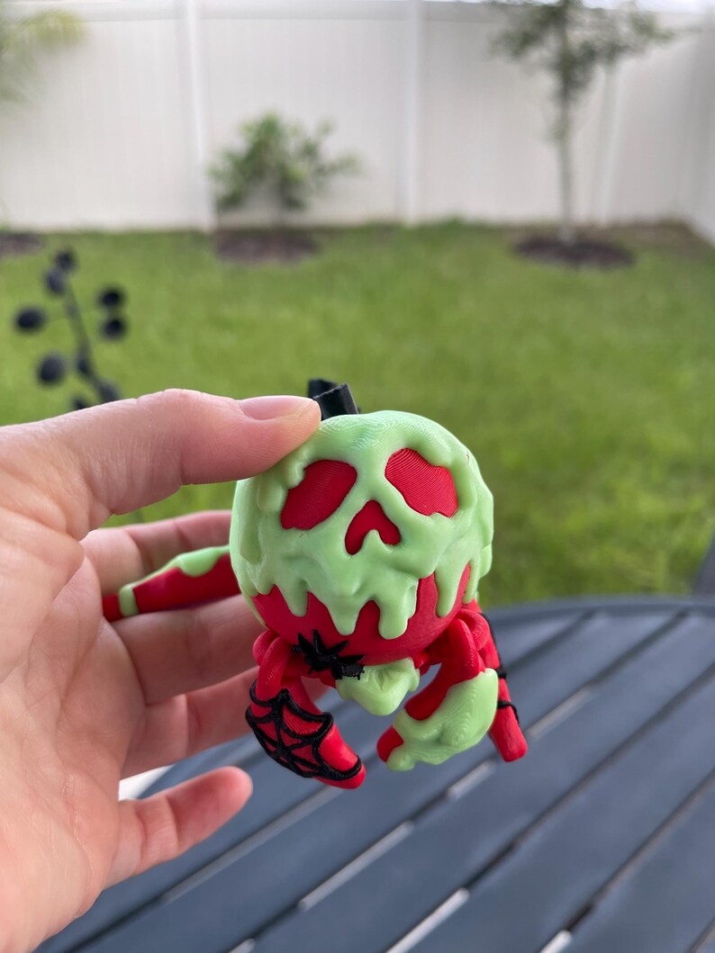 Puede incluir: Una figura de ara&ntilde;a roja y verde con tem&aacute;tica de Halloween. La ara&ntilde;a tiene un cuerpo rojo con detalles verdes goteando y detalles de telara&ntilde;a negros. La figura se sostiene en una mano sobre un fondo borroso al aire libre.
