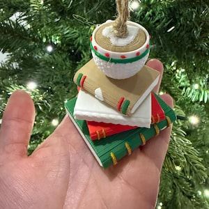K&ouml;nnte beinhalten: Ein Weihnachtsbaumschmuck in Form eines Stapels Miniaturb&uuml;cher, gekr&ouml;nt von einem runden, strukturierten Ornament. Die B&uuml;cher sind in Braun-, Wei&szlig;-, Rot- und Gr&uuml;nt&ouml;nen gehalten und weisen dekorative Details auf. Eine Schnurschlaufe ist zum Aufh&auml;ngen befestigt.